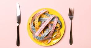 Top view of colorful measuring tapes on plate in the form of spaghetti with knife and fork on pink background.