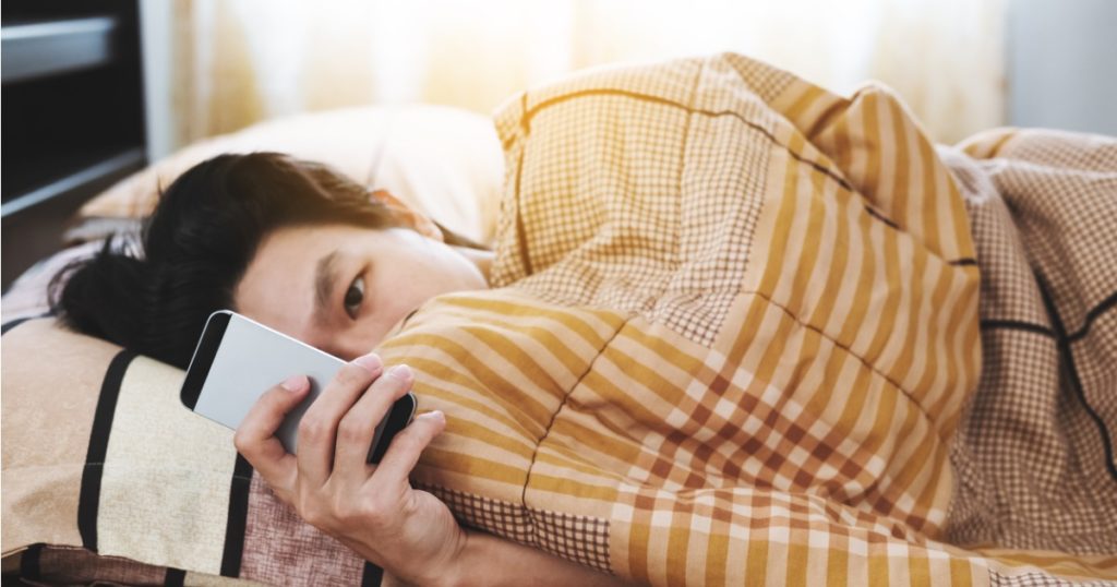 Person scrolling through their phone while laying under a blanket in bed.