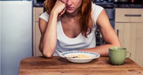 Closeup of woman sitting at the kitchen table, resting her cheek against a closed fist, staring at a bowl of cereal and a coffee mug.