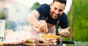 Man sprinkling salt on top of meats and vegetables cooking on an outdoor grill.