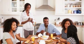 Mother, father and three teenage daughters laughing at the kitchen table over breakfast.