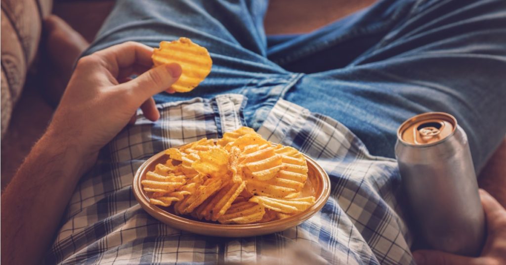Close-up of a man wearing a checkered shirt and jeans laying on his back with a plate of chips resting on his stomach and a can of beer in his right hand.