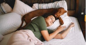 Photo of a woman sleeping in a bed, with a dog curled around her head.